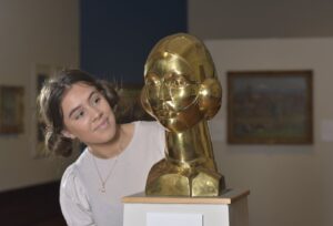 A young white woman with her hair in large buns in the side of her head next to the bronze statute Eastre – Hymn to the Sun