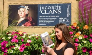 A young adult with light skein and long dark brown hair reading a paper programme underneath a sign advertising the Jacobite Clans exhibition at Perth Museum.