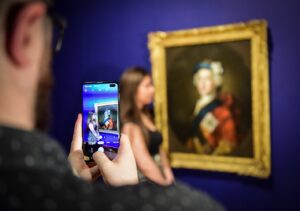 A light skinned adult taking a picture with his mobile phone of a young light skinned adult standing next to a painted portrait of Bonnie Prince Charlie at Perth Museum (Katie Adams and David McLeod)
