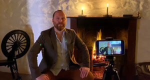 A man sits in front of a fire place with a mobile phone recording him. He's wearing a blue shirt and a brown jacket.