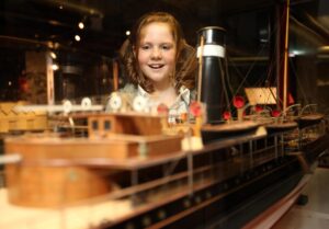 A child with light skin and red hair smiles as they look at a model ship.