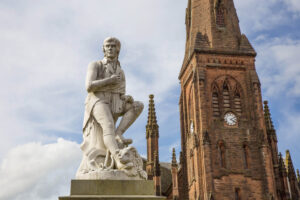 A white marble statue of Scottish poet, Robert Burns, stands in the foreground. A red sandstone church tower with a clock that is part of Greyfriars Kirk is in the background