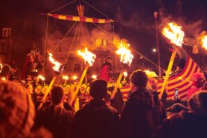 Crowds watch a torchlit procession at night. Two adults in Viking costume stand on board a Viking longship at the centre of the procession.