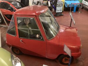 A young adult with light skin and wavy brown hair sits inside a small red car at Dundee Transport Museum.