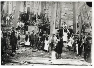 A black and white 19th century photograph of workers on a construction site. Dozens of adults and children stand between large wooden struts and unfinished stone walls.