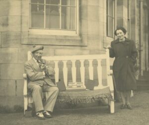 A sepia photograph of an older adult sitting on a bench. The older adult, who has light skin and is wearing a suit, is 20th-century Scottish comedian Sir Harry Lauder. Standing next to the bench is an adult with light skin, short wavy hair, and a long dark coat.