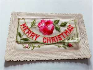 A white historical fabric and embroidered postcard, depicting embroidered pink flowers and green foliage. Embroidered text reads "Merry Christmas".