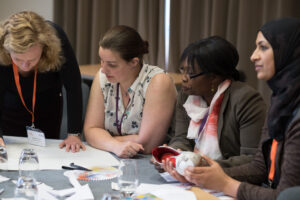 Two light skinned people with blonde and brown hair, sit next to a dark skinned person with short black hair in a ponytail and a medium skinned person wearing a dark hijab. They sit around a table looking at documents