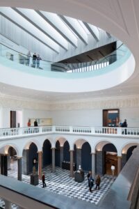 The entrance hall of Aberdeen Art Gallery. Two layers of balconies overlook a bright, open space with neo-classical pillars. Groups of visitors walk through the space.