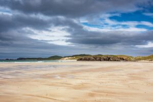 A wide sandy beach under a cloudy sky. There are low hills and sand dunes in the distance.