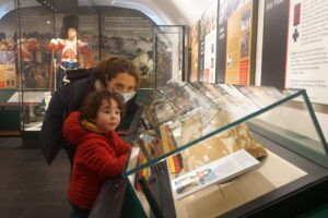 An adult with light skin, brown hair tied into a ponytail, and a face mask leans forward next to a child with light skin and curly brown hair. They are looking in a case containing military objects at the Argyll and Sutherland Highlanders Museum.