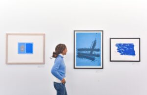 An adult with medium-dark skin, brown braided hair and a light blue jumper looks at art in the University of Stirling gallery. The artworks are all in various shades of blue.