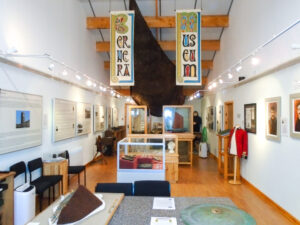 A museum gallery with white walls, a wooden floor, and a variety of display cases. A large wooden boat is suspended from the ceiling.