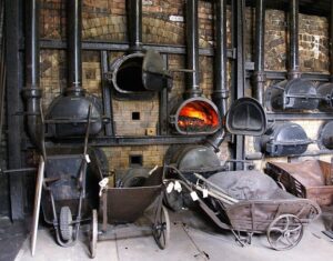 Wheelbarrows positioned in front of a row of black metal furnaces. An orange light glows from one of the furnaces.