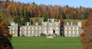 A large, long house made of grey stone. It has rows of tall white windows and a landscaped lawn. Behind the house is a forest of trees with yellow and orange leaves.