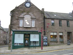 A two-storey building made of dark red stone. It has a large arched window, and a clock in its gable.