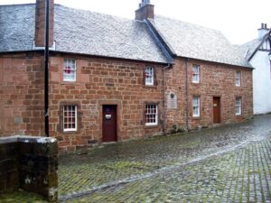 Two adjacent two-storey buildings with red stone walls, grey tiled rooves and small white windows. They are located on a sloping cobbled street.