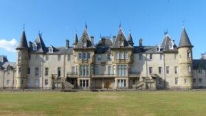 A large three storey stately home with sandy stone walls, pointed grey tile rooves, and gothic features.