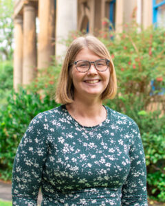 An adult with light skin, medium-length straight light brown hair, glasses, and a green floral dress.