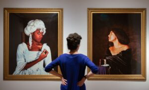 An adult with light skin and brown hair tied into a bun views a pair of portraits by Maud Sulter, a Scottish artist of Ghanaian heritage, at Edinburgh’s City Art Centre. One portrait shows an adult with medium-dark skin wearing a white wig and formal dress in the style of 18th century Western fashion. The other shows an adult with medium-tone skin wearing a black velvet robe.