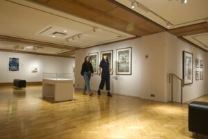Two adults with light skin walk through a gallery at the City Art Centre. The gallery has white walls and parquet flooring, and displays varied contemporary works.
