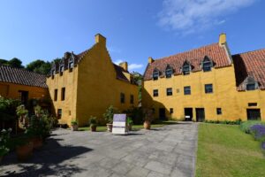 Two three-storey buildings with bright yellow walls, rows of small windows, and orange tiled rooves. Connecting the two buildings is a flagstone path with runs through a garden.