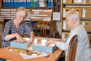 Two older adults with light skin sit at a table in Cupar Museum. They are going through a pile of documents and adding them to the museum’s collection.