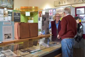 Two older adults with light skin have a conversation while standing in the display space at Cupar Museum. They are surrounded by documents, panels, and artefacts.