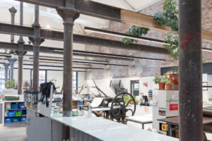 Printing presses, art supplies, and an array of pot plants fill a large workspace with brick walls and tall grey metal columns.