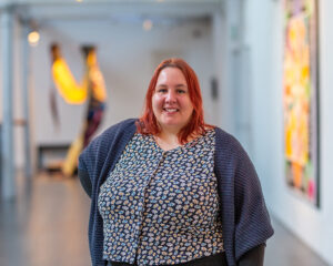 a light skinned person with shoulder length auburn hair, wearing a dotted shirt and a blue cardigan, stands with their right arm on their hip