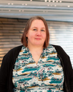 An adult with light skin, straight red shoulder-length hair, and a green and white patterned dress.