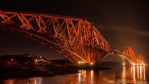 The Forth Railway Bridge, a large metal bridge which spans the Firth of Forth, illuminated against the night sky. It is formed of a complex web of orange-red metal struts and scaffolds.