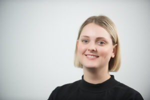 A headshot of a light skinned person with chin length bobbed blonde hair, wearing a black roll neck jumper