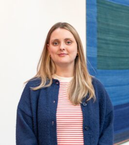 A headshot of a light skinned person with long straight blonde hair, wearing a dark blue cardigan and a red-and-white striped t-shirt.