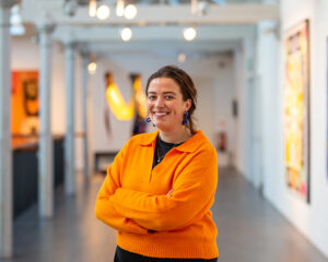 A light skinned person with waxy brown hair pulled back in a ponytail, wearing a bright orange jumper, stands with their arms crossed