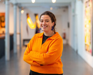 A light skinned person with waxy brown hair pulled back in a ponytail, wearing a bright orange jumper, stands with their arms crossed