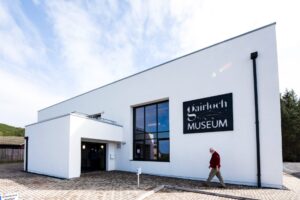 The entrance to Gairloch Museum, a minimalist building with a flat roof and white walls. An older adult with light skin and white hair is walking towards the front door.