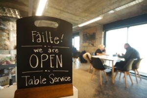 A small blackboard featuring the words 'Fàilte! We are open. Table Service' in white handwriting. In soft-focus in the background, two adults with light skin are sitting at a table.