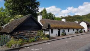 A long one-storey building with white walls and a thatched roof. Next to it is a building with rough stone and black wooden walls, and a thatched roof. There are several trees behind the buildings.