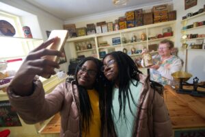 Twin teenagers with dark skin and long black braided hair stand and pose for a selfie in a mid-20th century sweet shop. An older adult with light skin and a floral apron stands and smiles behind the teenagers.