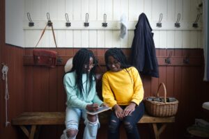 Two teenagers with dark skin and long black braided hair sit together in an early 20th-century school cloakroom. One teenager, who is wearing a mint green jumper, writes on a small slate. The other teenager, who is wearing a yellow jumper, smiles and watches.