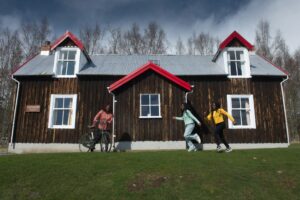 An adult with dark skin and medium-length red braided hair and twin teenagers with dark skin and long black braided hair play in front of a wooden building white a grey metal roof at the Highland Folk Museum. The teenagers are running towards the adult, who is standing with a bicycle.