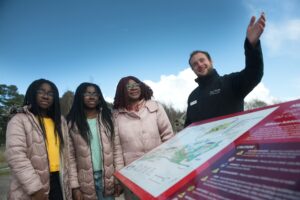 Two teenagers with dark skin and black braided hair, an adult with dark skin and red braided hair, and an adult with light skin and short brown hair stand together in front of a map panel. The adult with light skin and short brown hair gestures forward with their hand.