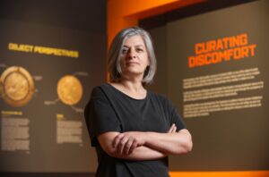 An adult with medium-light skin and grey shoulder-length hair stands with their arms folded in front of a display featuring the words 'Curating Discomfort' in orange text.