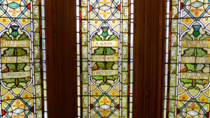 Three stained glass windows with elaborate floral designs. In the centre of each window is a design of a scroll wrapped around a thistle. Each scroll features an idiom written in the Scots language.