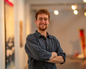 A light skinned person with short brown curly hair and a brown goatee beard, wearing a grey shirt, stands with their arms crossed