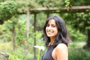 An adult with medium skin tone and shoulder length dark hair. They are smiling at the camera. In the background there is a green garden area.