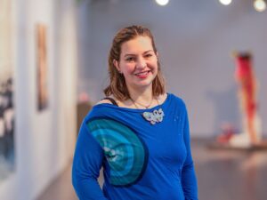 A light skinned person with shoulder length brown hair, partial tied back in a ponytail, wearing a bright blue top with a mandala pattern on it, stands with their hands in their pockets