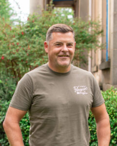 An adult with medium-light skin, short brown hair and a brown t-shirt, standing in a garden.