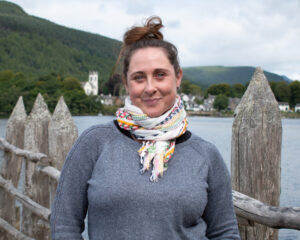 An adult with light skin and brown hair tied into a bun. They are wearing a grey jumper and a multicoloured scarf.